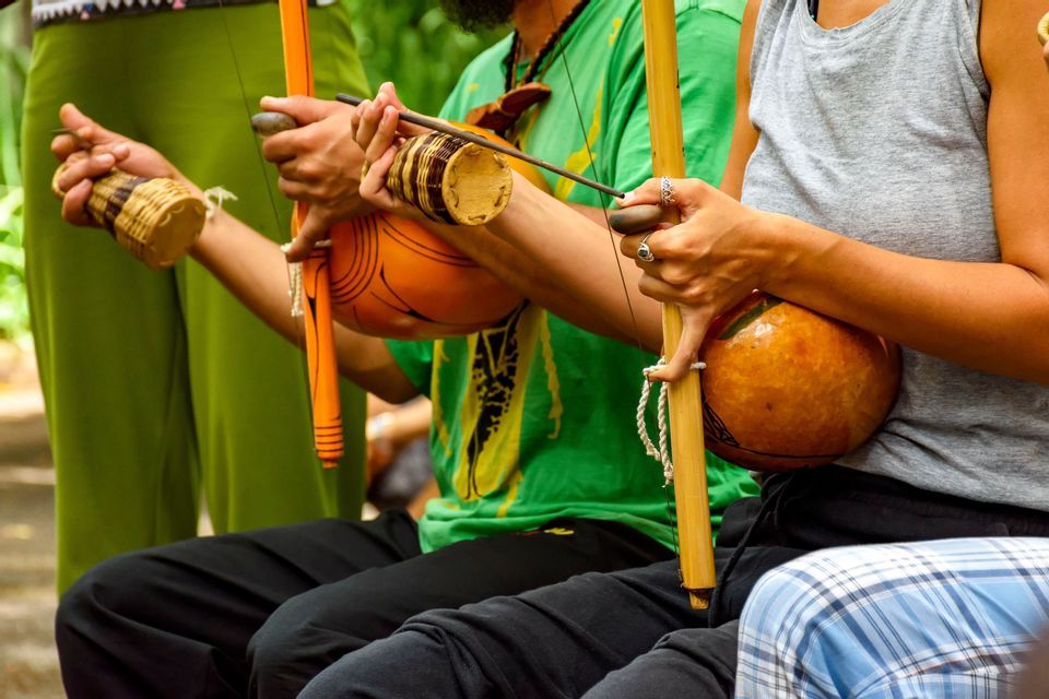 Un primer plano de tres personas sentadas sosteniendo instrumentos musicales tradicionales de berimbau y un agitador caxixi.