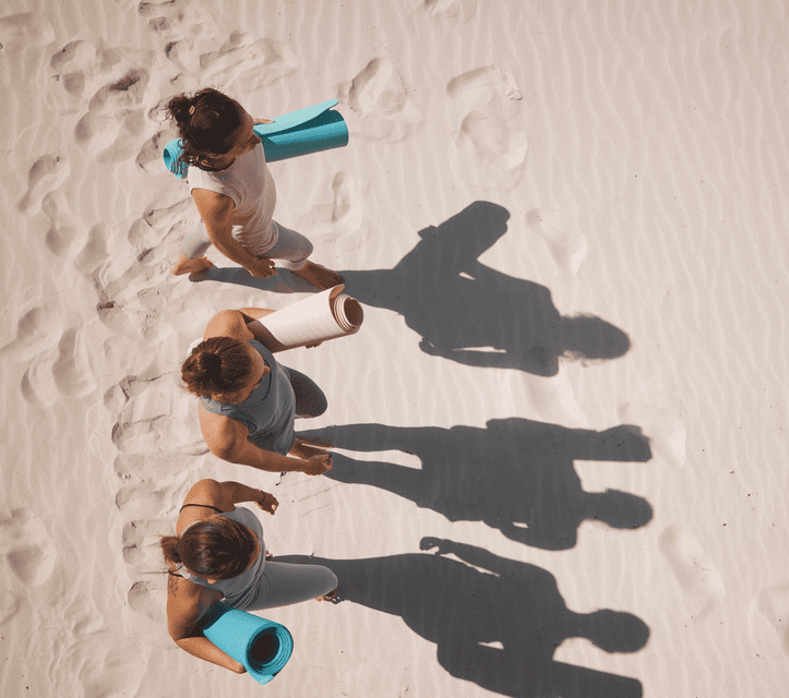 Luftaufnahme von drei Personen einer WeRoad-Gruppenreise, die im Sand gehen, zusammengerollte Yogamatten tragen und lange Schatten werfen.