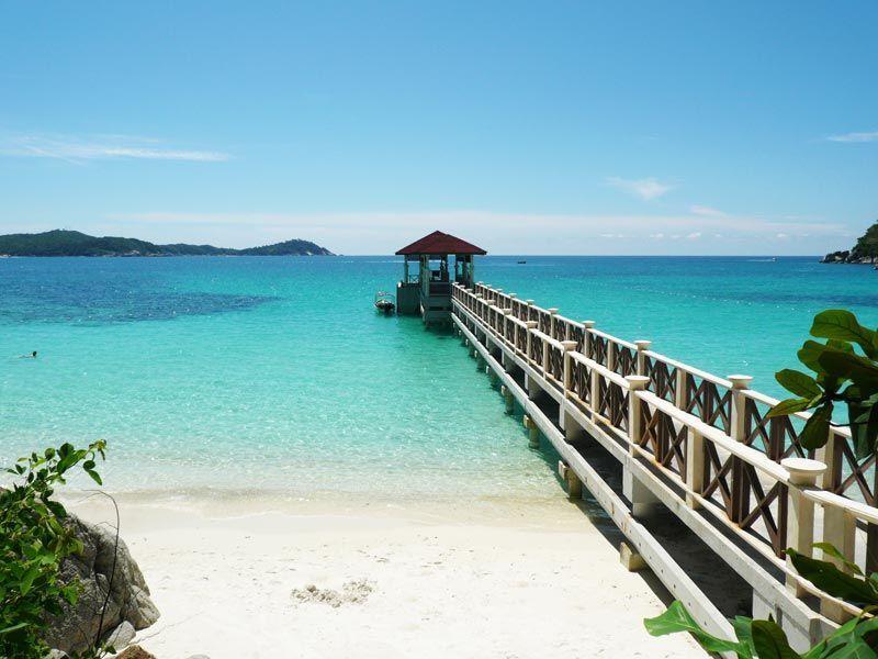 Un pontile di legno con un gazebo si estende da una spiaggia di sabbia bianca su acque turchesi limpide verso isole verdi e lontane.