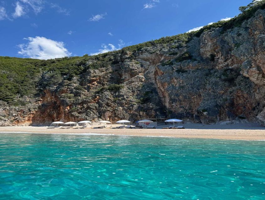 Una spiaggia sabbiosa con ombrelloni bianchi e lettini si trova alla base di un'ampia scogliera rocciosa, vista dall'acqua turchese.