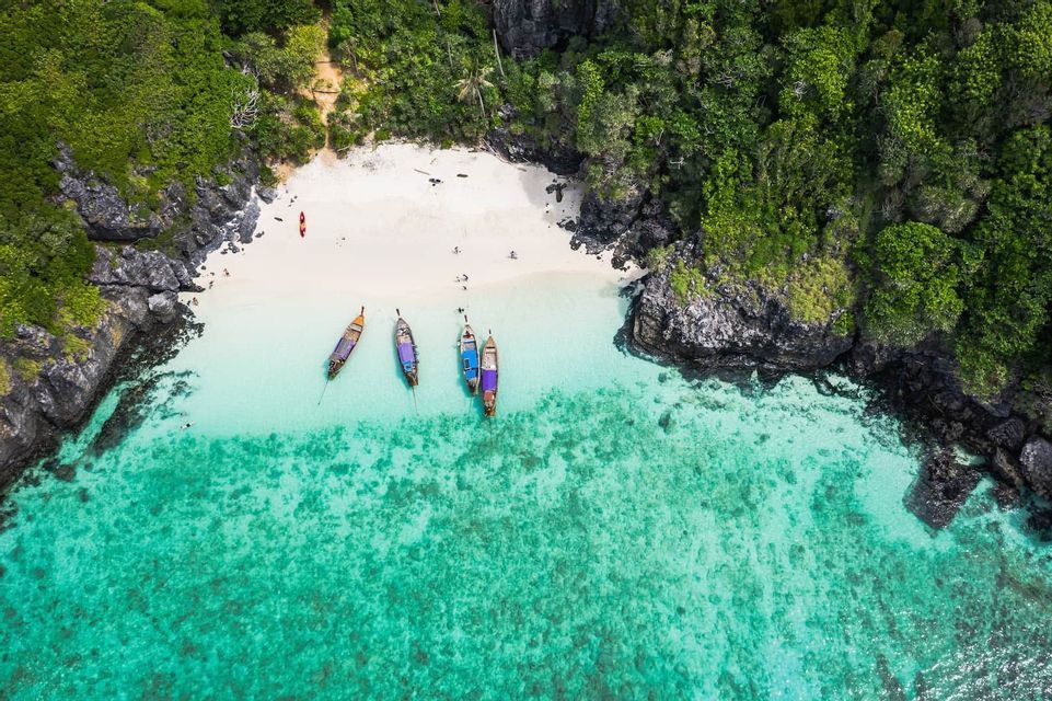 Una vista aérea de varios barcos tradicionales amarrados en agua cristalina color turquesa junto a una playa de arena blanca aislada rodeada de exuberantes acantilados verdes.