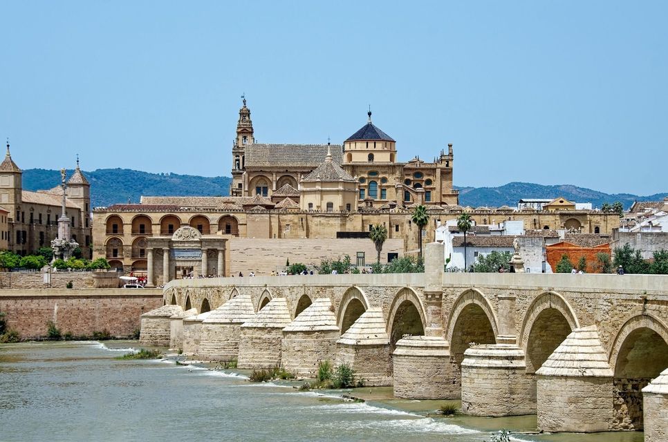 Un ponte di pietra a più arcate attraversa un ampio fiume, conducendo a una città storica con una grande cattedrale e un campanile.