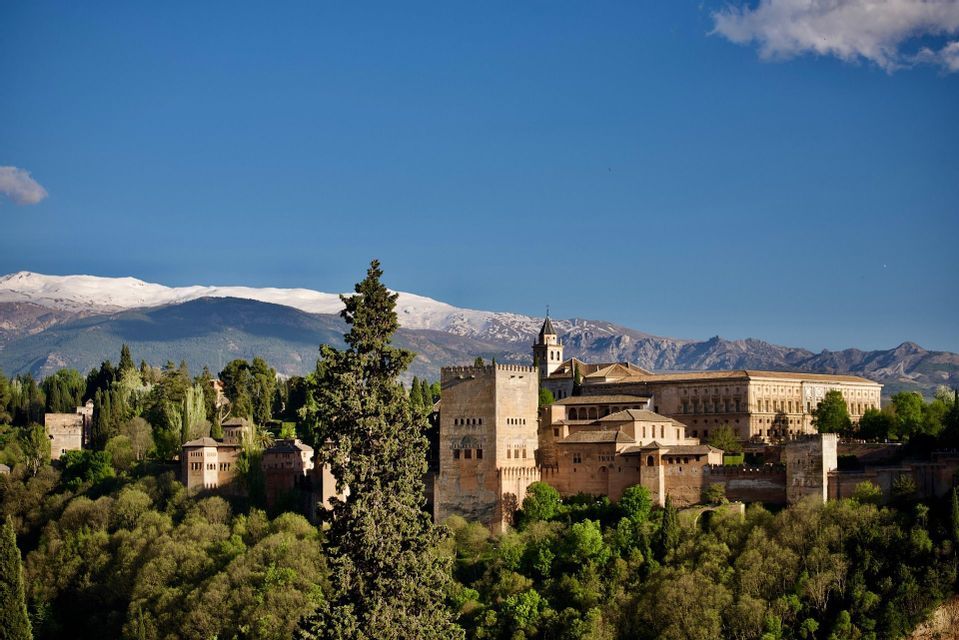 Un complesso storico di palazzi sorge su una verde collina, con una catena montuosa innevata sullo sfondo e un cielo azzurro e limpido.