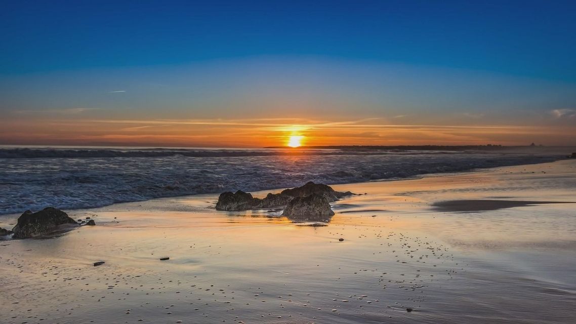 Il sole tramonta sull'oceano, la sua luce si riflette sulla sabbia bagnata di una spiaggia con scogli nell'acqua bassa.