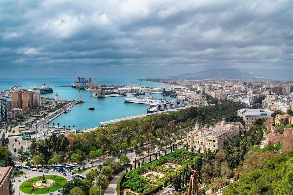Vista aerea di un porto costiero, con navi da crociera e parchi verdi rigogliosi in primo piano, sotto un cielo nuvoloso.