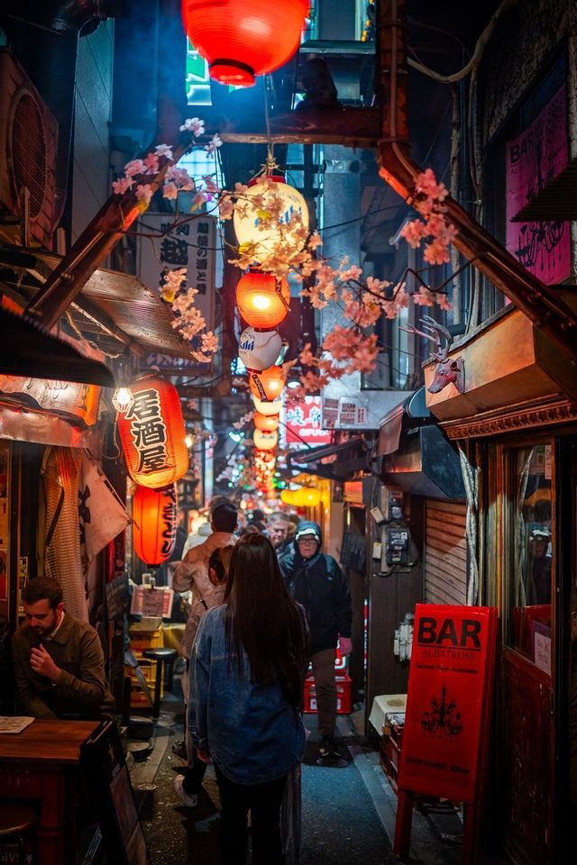 Una folla di persone cammina lungo un vicolo stretto di notte, illuminata da sfavillanti lanterne di carta rosse e bianche e fiori di ciliegio.
