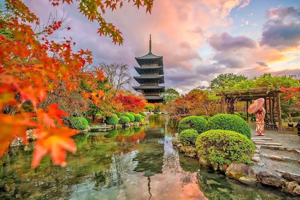 Una pagoda giapponese si specchia in uno stagno, con una persona in kimono tra il vibrante fogliame autunnale.
