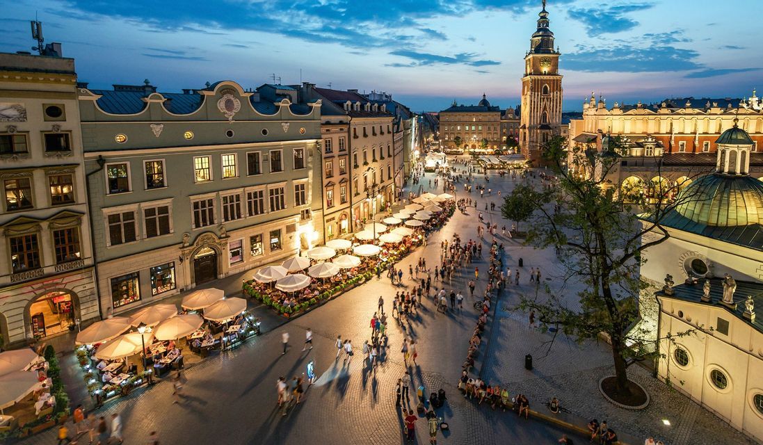 Una vista dall'alto di una vivace piazza storica al tramonto, piena di gente che passeggia e cena ai caffè all'aperto sotto gli ombrelloni.