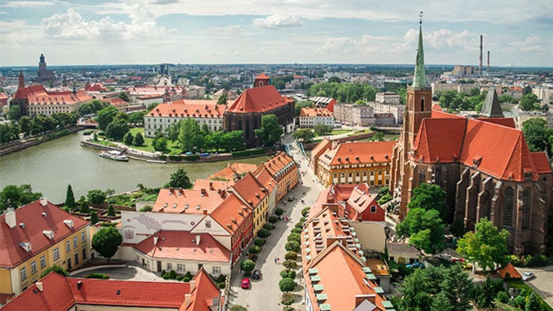 Una vista aerea di una storica città europea con tetti in tegole rosse, una chiesa imponente e un fiume che la attraversa in una giornata di sole.