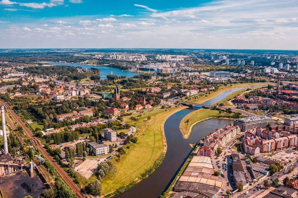 Una vista aerea di una città tentacolare dove un fiume scorre tra rigogliose aree verdi e gruppi di edifici sotto un cielo blu.