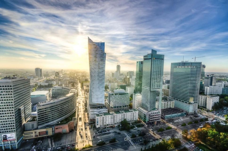 Una vista aerea dello skyline di una città moderna con numerosi grattacieli e strade trafficate sotto un cielo luminoso e soleggiato.