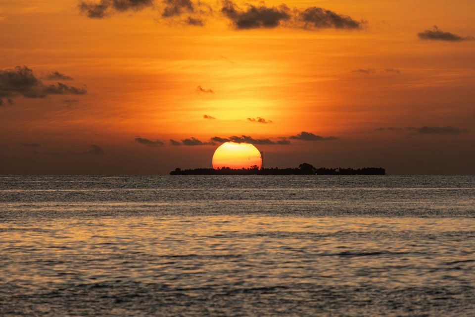 Un grande sole arancione tramonta sull'oceano, parzialmente nascosto da un'isola in silhouette sotto un cielo nuvoloso.