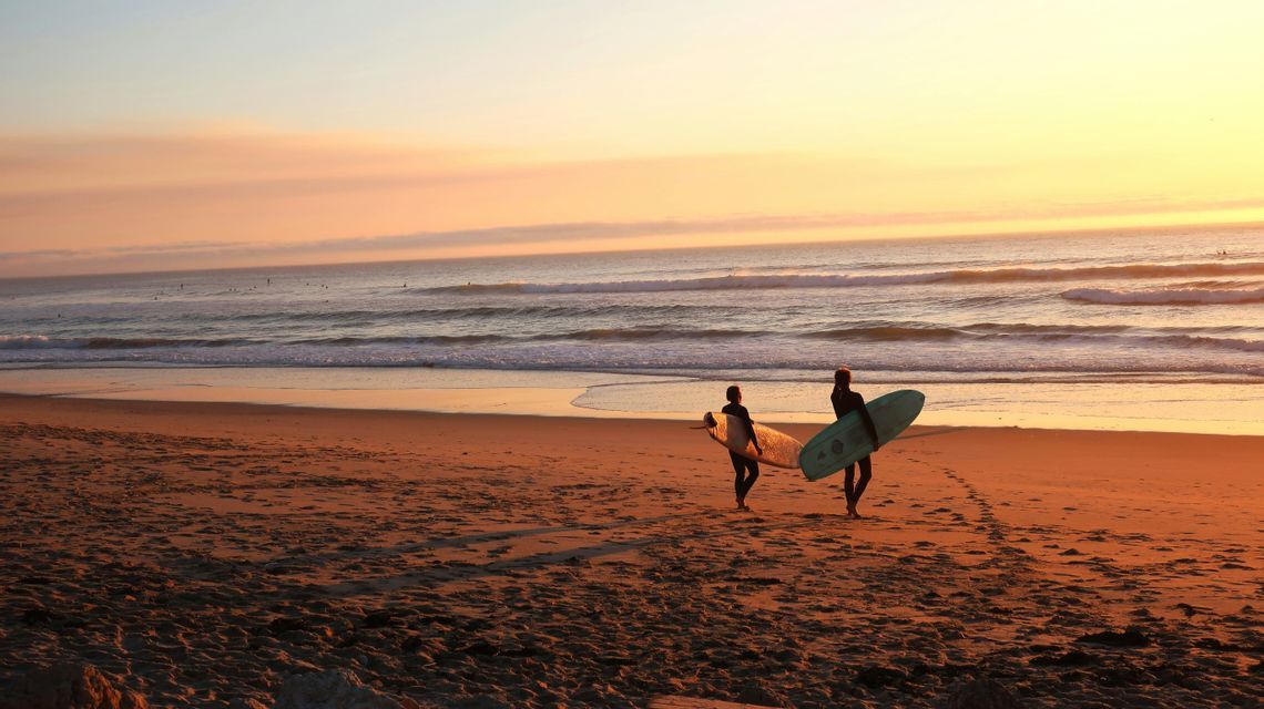 Due surfisti in muta portano le tavole da surf camminando lungo una spiaggia sabbiosa verso l'oceano, al tramonto caldo.