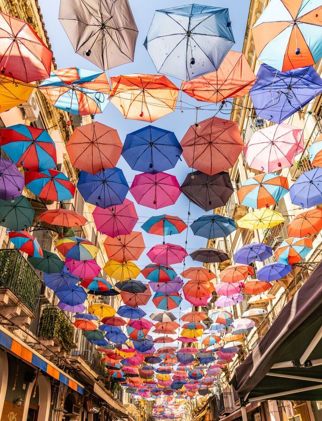 Una veduta dal basso di centinaia di ombrelli colorati sospesi sopra una via stretta tra gli edifici, sotto un cielo blu.