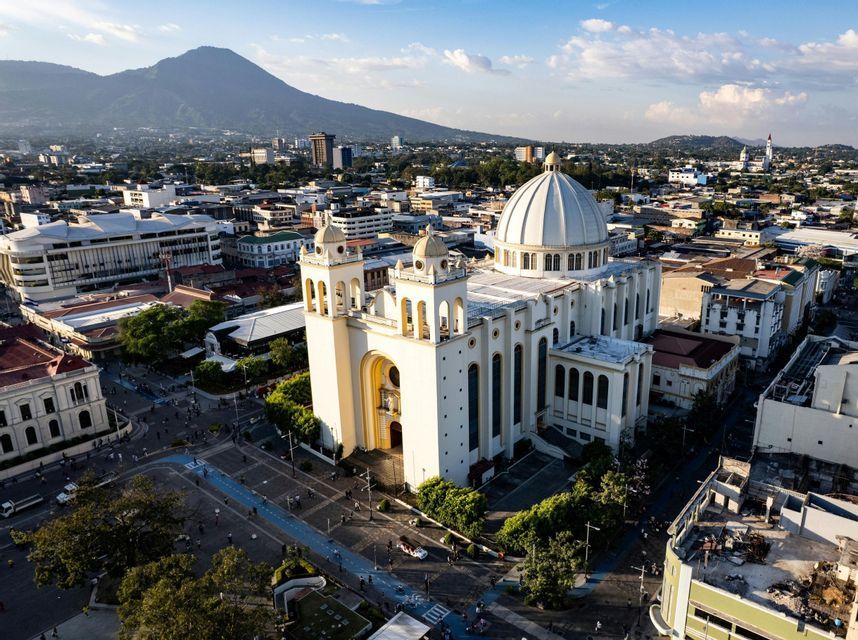 Veduta aerea di una grande cattedrale bianca con cupola, al centro di una città e un vulcano sullo sfondo.
