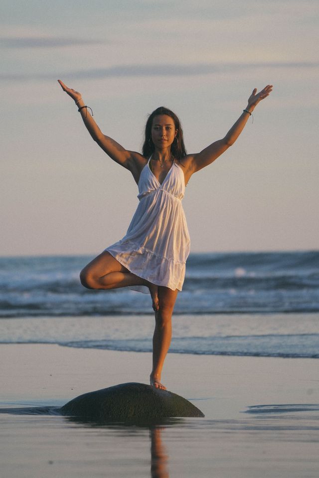 Una donna in abito bianco pratica la posizione dell'albero yoga in equilibrio su una roccia su una spiaggia al tramonto.