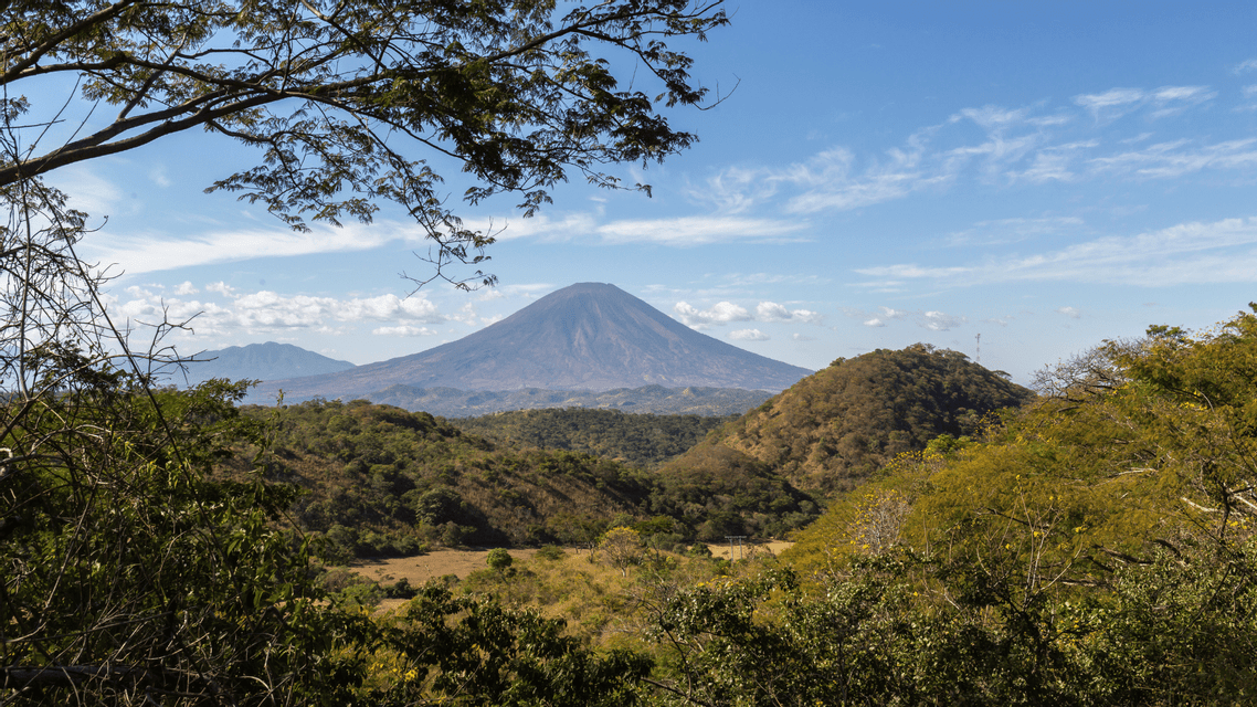 Un grande vulcano conico si erge in lontananza su una valle verde lussureggiante e dolci colline, incorniciato da rami d'albero.