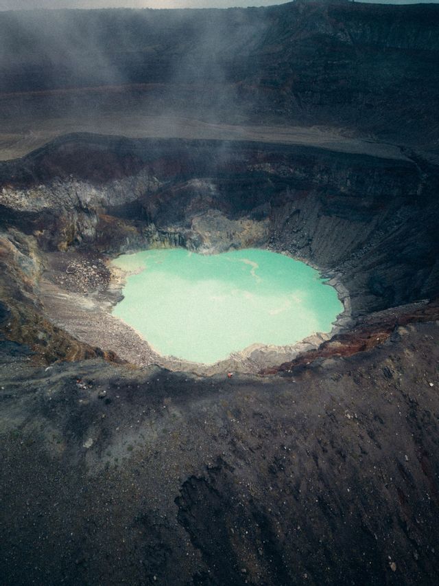 Veduta aerea di un lago turchese brillante nel fondo di un vasto e scuro cratere vulcanico, con vapore che sale dalle pendici.