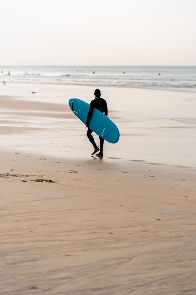 Una persona in muta nera cammina lungo una spiaggia sabbiosa, trasportando una tavola da surf blu verso l'oceano.