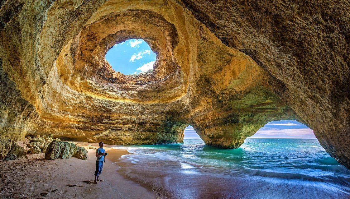 Una persona si trova su una spiaggia sabbiosa all'interno di una grande grotta marina con un oculo nel soffitto e un arco che si apre verso il mare.