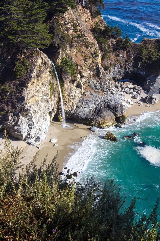 Una cascata scende da una scogliera alberata su una spiaggia sabbiosa che si affaccia su un mare turchese.