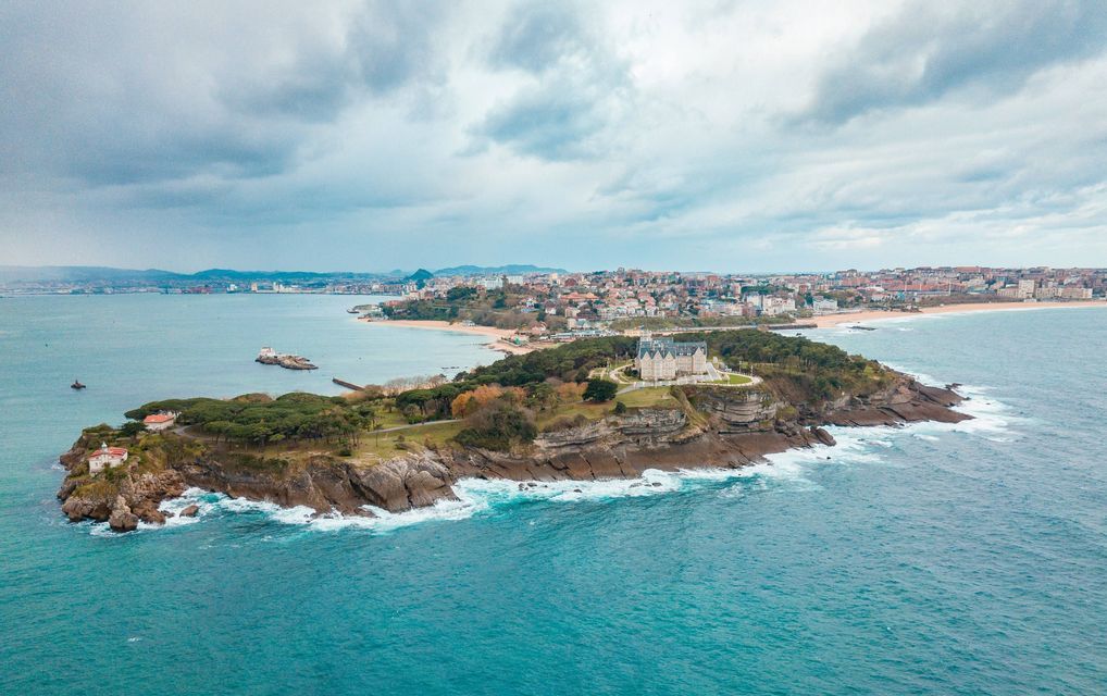 Veduta aerea di un palazzo su una penisola boscosa che si protende nell'oceano, con una città costiera sullo sfondo sotto un cielo nuvoloso.