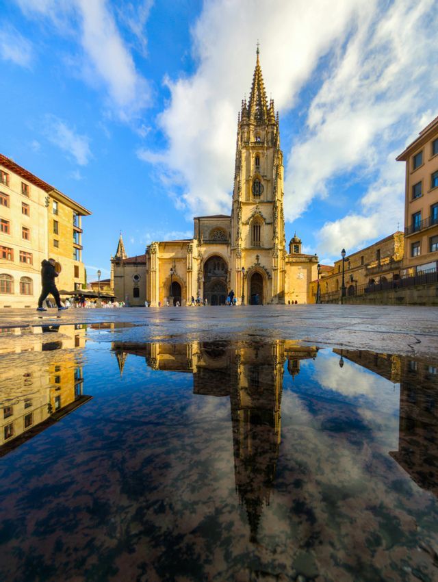 Una vista dal basso di una cattedrale gotica con un'alta guglia riflessa in una grande pozzanghera su una piazza di pietra.