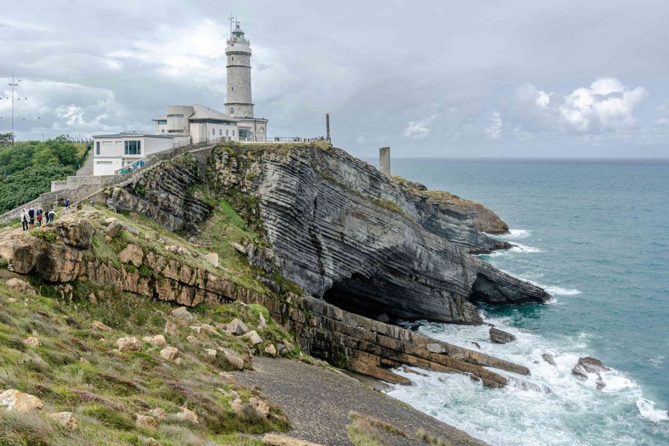 Un faro si erge su una scogliera rocciosa stratificata a picco sul mare, con un viaggio di gruppo WeRoad visibile su un sentiero lungo il fianco della collina.