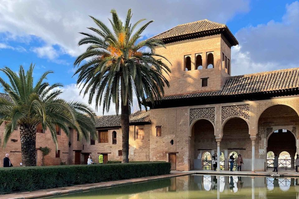 A brick building with arches and a tower next to a reflecting pool and palm trees in a courtyard.