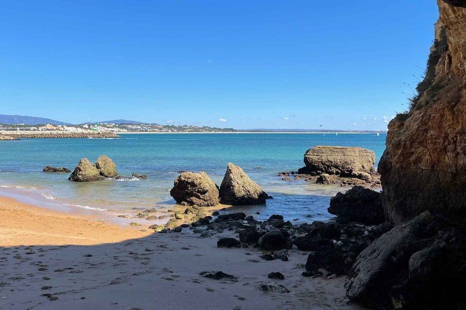 A view from a coastal cave looking out onto a sandy beach with large rocks in the turquoise sea under a clear blue sky.