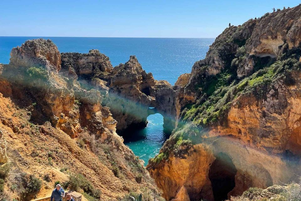 Un panorama da un sentiero a picco sul mare, con faraglioni dorati e un arco naturale su acqua turchese, e persone che passeggiano.