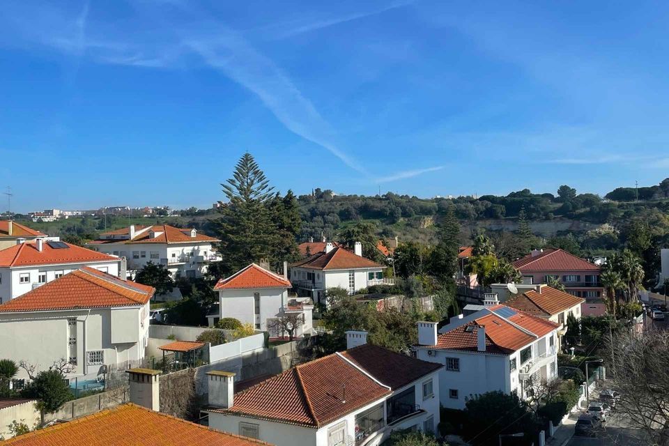 An aerial view of white houses with orange-tiled roofs scattered across a sunlit, green hillside under a clear blue sky.