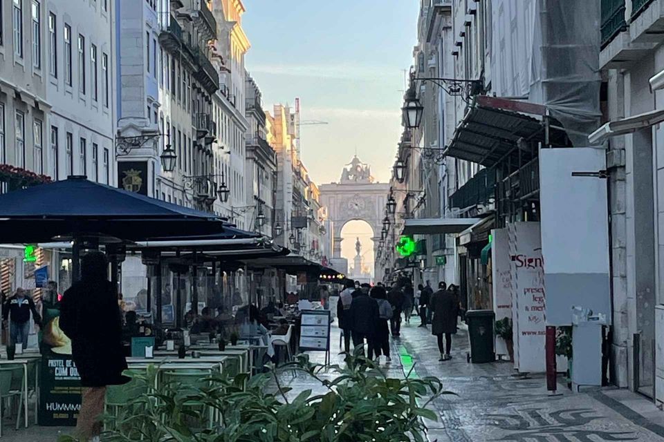 A pedestrian street lined with outdoor cafes and people walking towards a large triumphal arch in the distance.