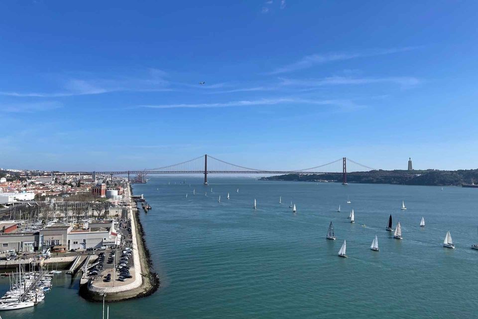A panoramic view of sailboats on a wide river next to a city marina, with a large red suspension bridge in the background.
