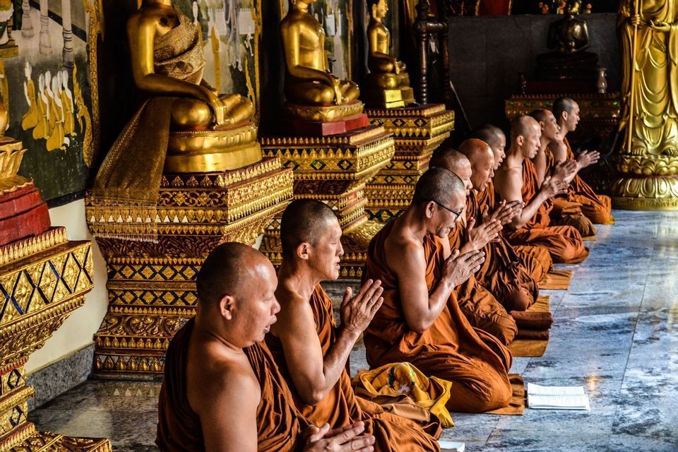 Eine Reihe buddhistischer Mönche in orangefarbenen Gewändern kniet betend in einem Tempel, mit goldenen Buddha-Statuen im Hintergrund.