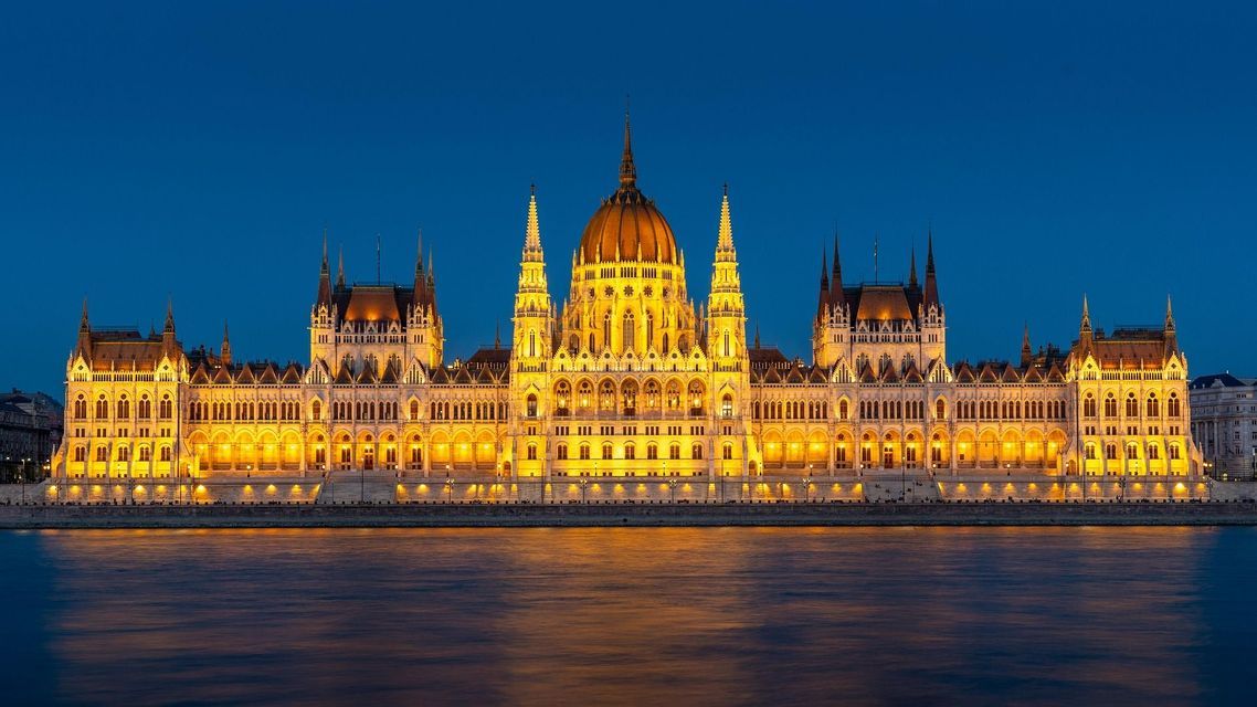 Un grande edificio sontuoso illuminato da luci dorate, che si riflette in un fiume al tramonto sotto un cielo blu intenso.