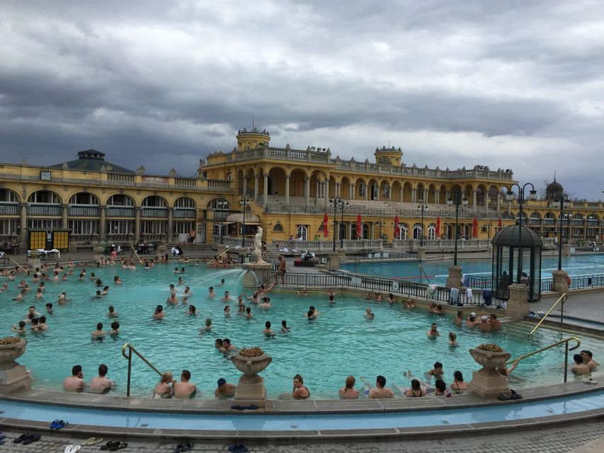 Molte persone si rilassano in una grande piscina termale all'aperto di fronte a un imponente edificio giallo sotto un cielo nuvoloso.