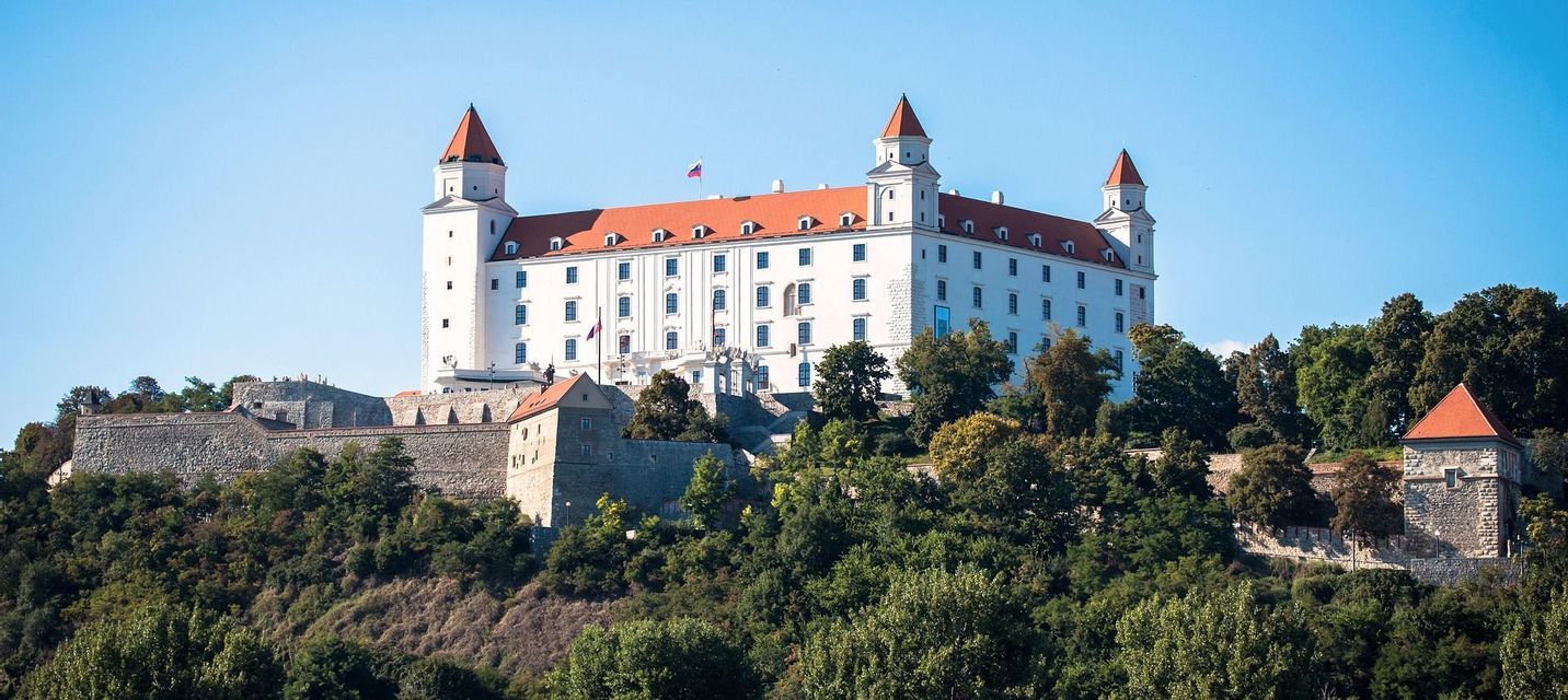 Un grande castello bianco con tetti rossi e quattro torri angolari si erge su una collina lussureggiante e alberata sotto un cielo azzurro e limpido.