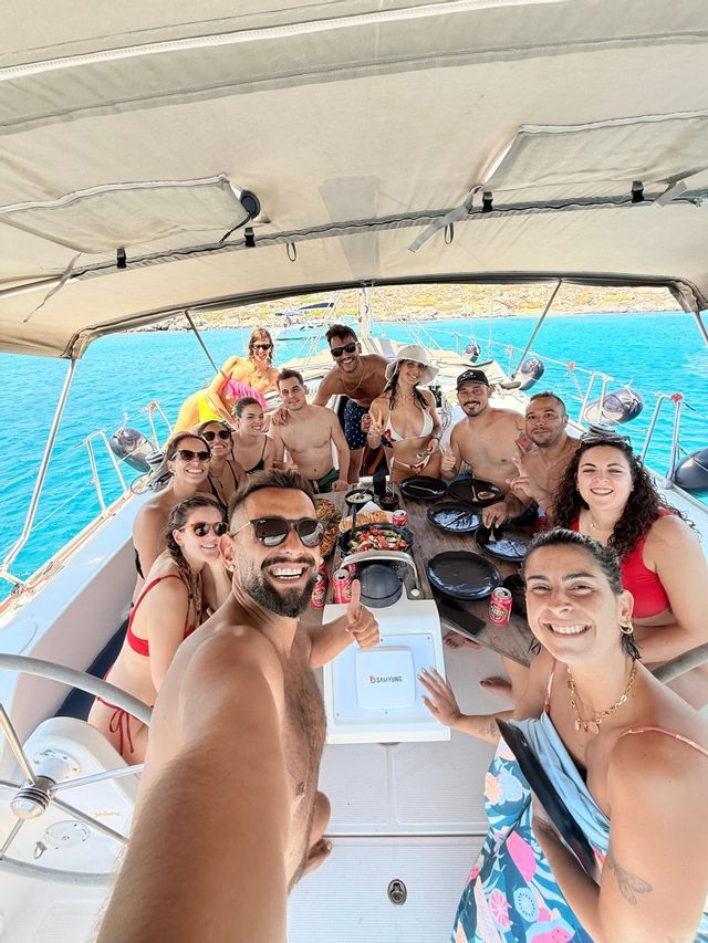 Un viaggio di gruppo WeRoad si scatta un selfie su una barca a vela, riuniti intorno a un tavolo con cibo e bevande in mare aperto.