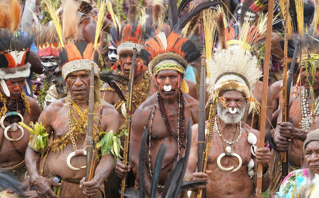 Un grupo de hombres indígenas con atuendos tradicionales, tocados de plumas, pintura corporal y joyas, portando lanzas.