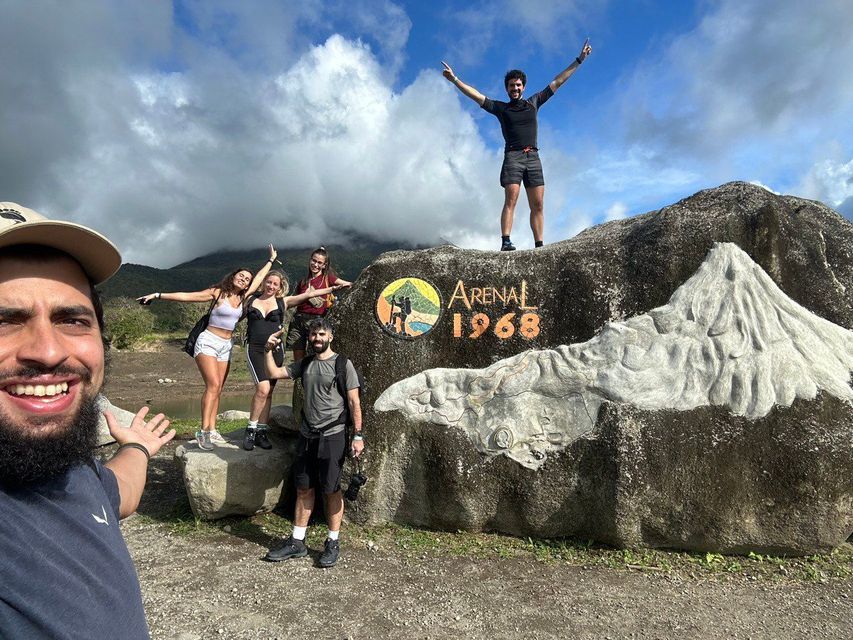 Un viaje en grupo de WeRoad se toma una selfie frente a una gran roca tallada con 'Arenal 1968' y un relieve de volcán.