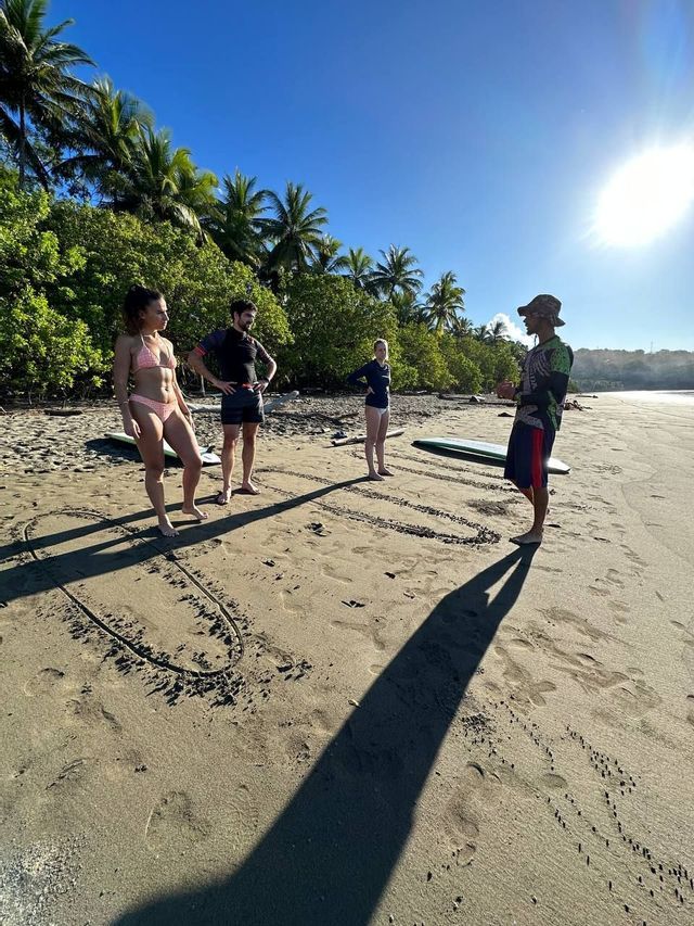 Un instructor imparte una lección de surf a un viaje en grupo de WeRoad en una playa soleada, con diagramas dibujados en la arena.