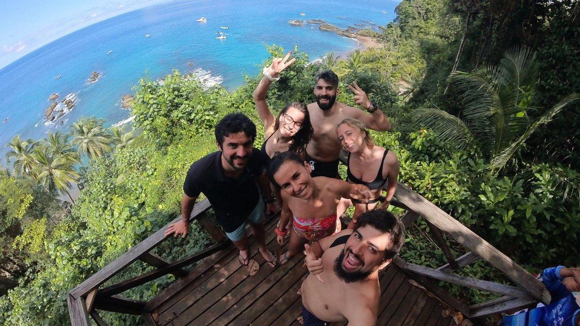 Un viaje en grupo de WeRoad se toma una selfie sonriente en un mirador de madera con vistas a una costa tropical de aguas azules.