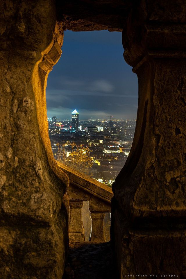 Skyline cittadino illuminato di notte, visto attraverso un arco in pietra dalla luce calda.