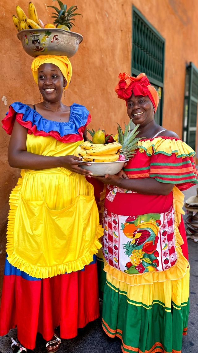 Zwei Frauen in farbenfrohen Trachten lächeln und balancieren Schalen mit Bananen und Ananas.