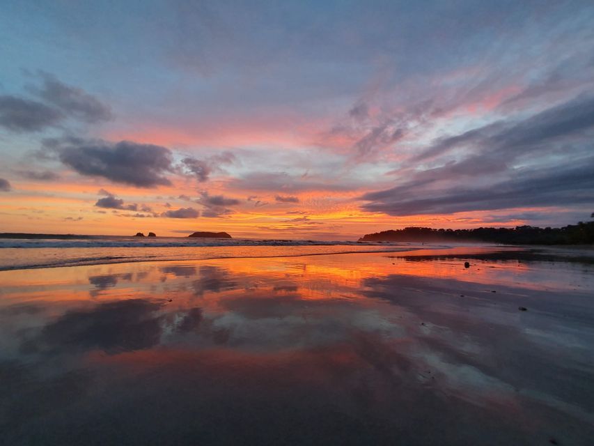 Un atardecer colorido con nubes naranjas y rosas se refleja en la arena mojada de una playa, con pequeñas islas en el horizonte.