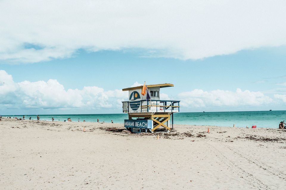 Una colorata torre di salvataggio etichettata 'Miami Beach' si erge su una spiaggia sabbiosa con l'oceano turchese e un cielo parzialmente nuvoloso sullo sfondo.