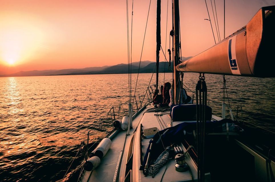Eine WeRoad-Gruppenreise an Deck eines Segelboots, die einen orangefarbenen Sonnenuntergang über dem Wasser mit fernen Bergen genießt.