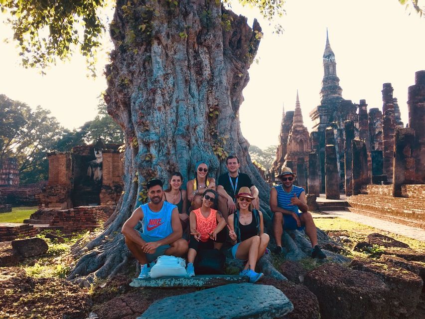 A WeRoad group trip poses for a photo at the base of a large, ancient tree with temple ruins in the background during sunset.