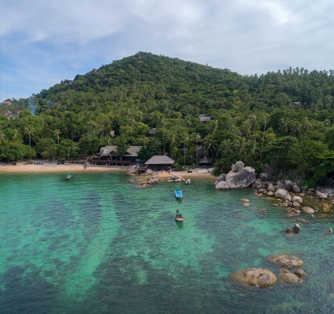 An aerial view of a tropical cove with clear turquoise water, small boats, a sandy beach, and a lush green, tree-covered hill.